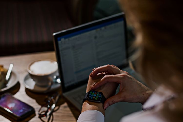 Woman looking at a smart watch in a coffee shop with a laptop, mobile phone, cake and coffee on the table